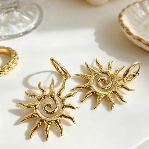 May include: A pair of gold-colored sunburst earrings with a spiral center. The earrings have a textured, hammered appearance and a hoop closure. The design is reminiscent of a sun with rays extending outwards.