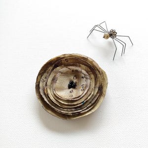 May include: A handmade, layered, circular brooch with a textured, earthy appearance. The brooch is accompanied by a small, metal spider with delicate legs and a decorative body. The brooch has a dark center with small black beads.