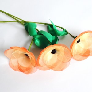 May include: Three peach-colored silk flowers with black centers on green stems. The flowers are made of sheer fabric and have a delicate, feminine look.