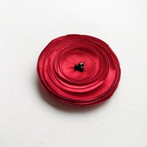 May include: A vibrant red fabric flower with layered petals, centered with three small black beads. The flower is circular and appears to be handmade, set against a white background. This decorative item could be used as a brooch or embellishment.