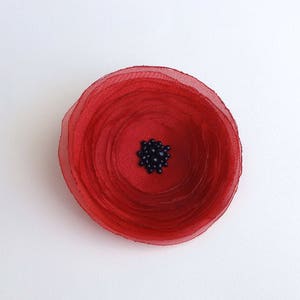 May include: A handmade red fabric flower with a dark center. The flower is circular with layered petals, and the center is filled with small, dark beads. The flower is set against a white background.