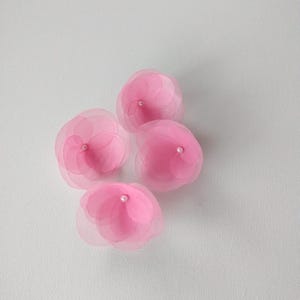 May include: Four delicate pink fabric flowers with a sheer, layered petal design. Each flower features a small, white bead at its centre. The flowers are arranged on a white background, creating a soft, feminine aesthetic.