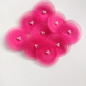 May include: A cluster of nine bright pink, layered fabric flowers with small white bead centers. The flowers are arranged in a slightly overlapping pattern against a white background. The petals have a translucent quality.