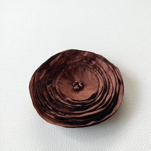May include: A circular fabric flower brooch in a deep chocolate brown color. The accessory features multiple layered, ruffled petals arranged in a concentric pattern, with three small, dark metallic beads clustered at the center of the piece.