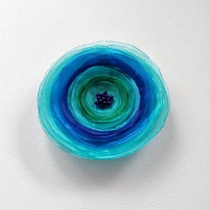 May include: A circular fabric flower brooch featuring layered, translucent petals in shades of turquoise, teal, and deep blue. The center of the flower is embellished with a cluster of small, dark blue beads, creating a textured, multi-dimensional appearance.