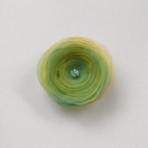 May include: A handmade fabric flower with layered petals in shades of green, yellow, and blue. The center of the flower features a small, sparkling blue gem. The flower is a decorative item, possibly for crafts or embellishment.