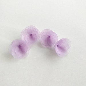 May include: Four lavender-coloured fabric flower embellishments. Each flower is made of multiple layers of sheer fabric, creating a delicate and layered look.