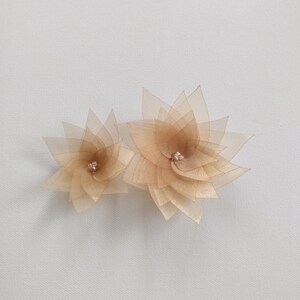 May include: Two delicate, layered flower hair accessories in shades of beige and tan. Each flower features multiple petals and a small, sparkling rhinestone center. The accessories are designed for formal occasions.