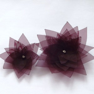 May include: Two burgundy fabric flowers with a layered, starburst design. The flowers have a small, clear rhinestone in the center.