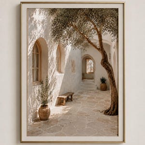 May include: A framed wall art print depicting a sunlit Mediterranean courtyard with white stone walls, arched windows, a stone-paved floor, a wooden bench, two potted plants, and a large tree with branches casting shadows across the scene.