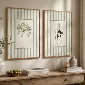 May include: Two framed wall art prints featuring botanical illustrations of wildflowers centered on a cream background with vertical sage green stripes. Each rectangular print is encased in a light wood frame, displaying detailed floral sketches.