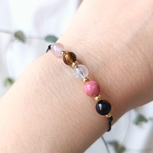 Peut inclure: Un bracelet de perles avec des pierres précieuses roses, marron, transparentes et noires, séparées par des entretoises dorées. Le bracelet est monté sur un cordon noir. Le bracelet est présenté sur un poignet.