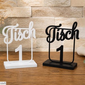 Könnte beinhalten: Zwei Tischnummern, eine in Weiß und eine in Schwarz, die jeweils das Wort Tisch in Schreibschrift über der Ziffer 1 zeigen. Die Schilder sind auf rechteckigen Sockeln montiert und haben ein schmales, aufrechtes Rahmendesign.