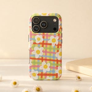 May include: A smartphone case featuring a colourful plaid pattern in shades of pink, orange, yellow, and blue, overlaid with white daisy flowers with yellow centres. The case has a black camera module cutout at the top with three circular lenses.