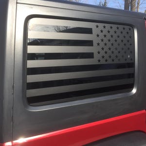 Könnte beinhalten: Schwarzer amerikanischer Flaggen-Fensteraufkleber auf einem roten Jeep. Das Flaggen-Design zeigt schwarze Streifen und Sterne und sorgt für einen patriotischen Look. Der Aufkleber wird auf der Heckscheibe des Fahrzeugs angebracht.