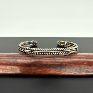May include: A silver and gold-toned cuff bracelet with a twisted rope design. The bracelet is displayed on a dark wooden surface against a black and white background. The cuff is approximately 2.5 inches in diameter.