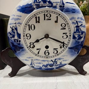 Op de afbeelding: Een ronde keramische klok met een blauw en wit Hollands windmolenontwerp. De wijzerplaat heeft zwarte cijfers en wijzers. Hij wordt gepresenteerd op een donkerbruine houten standaard. De klok is gemarkeerd met "Germany".