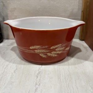 May include: A vintage red Pyrex bowl with a white interior and two handles. The exterior features a wheat pattern in a cream color. This kitchenware item is ideal for serving or storage.