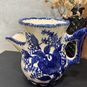 May include: A cream and blue ceramic shaving scuttle with a floral pattern. The scuttle features a blue floral design on a cream base, a blue handle, and a scalloped, blue-trimmed rim. It has a spout on the side.
