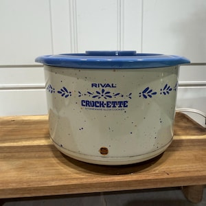 May include: A Rival Crock-Ette stoneware slow cooker with a cream-colored body featuring blue speckles and a blue floral pattern. The front displays the text Rival Crock-Ette Stoneware Slow Cooker and includes a small orange power indicator light.