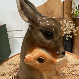 May include: A glossy ceramic vase shaped like the heads of a large deer and a small fawn nestled together. The adult deer is dark brown with large black eyes, while the fawn is a lighter tan color. The vase features a smooth, reflective finish.