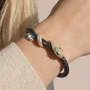 May include: A braided black leather bracelet featuring a gold-toned snake design that wraps around the band. The snake is embellished with clear sparkling crystals and has two small green stones set as eyes on the head of the serpent.