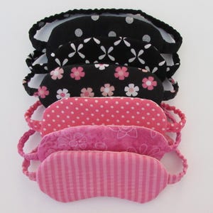 May include: A stack of six sleep masks. The masks are made of fabric and have different patterns. The top mask is black with white polka dots, the second mask is black with a white geometric pattern, the third mask is black with white flowers, the fourth mask is pink with white polka dots, the fifth mask is pink with a floral pattern, and the bottom mask is pink with white stripes.