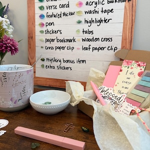 May include: A Mystery Blessings sign lists items including a bracelet, verse card, sticker, pen, notebook, bookmark, washi tape, highlighter, tabs, cross, and paper clips. A box contains stationery, a mug, a bowl, and small green leaf charms.