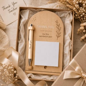 Pode incluir: Um quadro de notas de madeira clara em forma de arco com um suporte para caneta e uma esferográfica. O quadro apresenta o texto gravado "THANK YOU For this wonderful year" e uma ilustração de folha. Um bloco de notas branco quadrado está fixado no centro.
