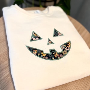 May include: A cream-colored crewneck sweatshirt featuring a jack-o'-lantern face design. The eyes, nose, and mouth are appliquéd with a dark fabric featuring a colorful floral pattern of yellow, pink, and blue flowers, outlined in green stitching.