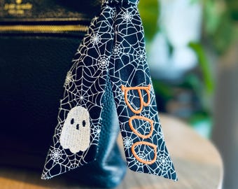 May include: A black fabric bag charm featuring a white spiderweb pattern, an embroidered white ghost, and the orange embroidered text BOO. The charm is tied to the handle of a black textured leather handbag with a gold zipper and logo hardware.