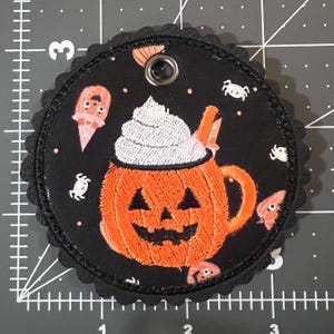 May include: A circular black fabric patch with a scalloped edge featuring an embroidered orange jack-o'-lantern mug topped with white whipped cream. The background pattern includes small white spiders and pink ghost figures. A metal grommet is at the top.