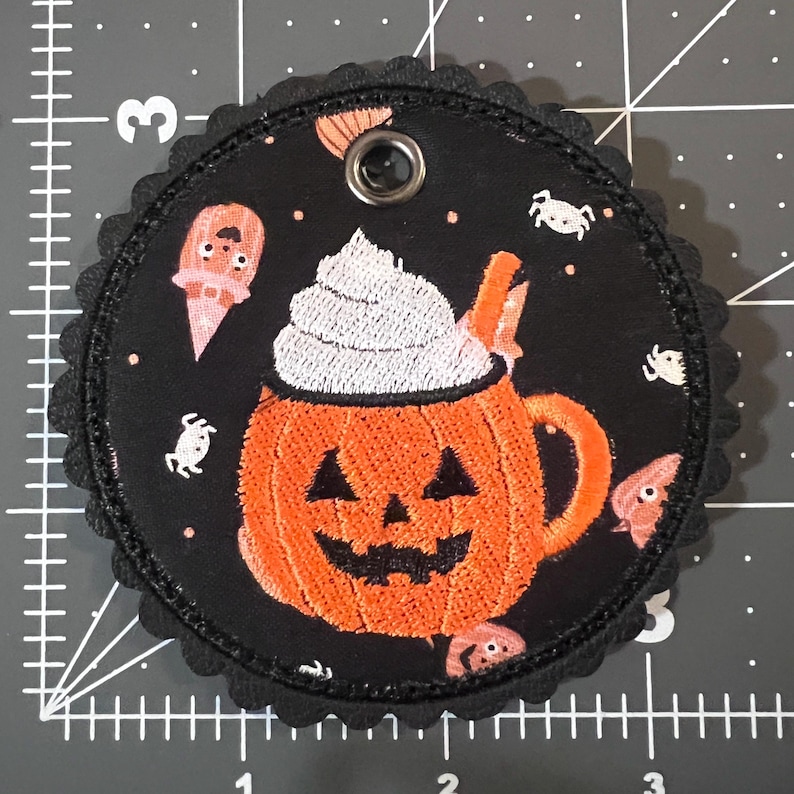 May include: A circular black fabric patch with a scalloped edge featuring an embroidered orange jack-o'-lantern mug topped with white whipped cream. The background pattern includes small white spiders and pink ghost figures. A metal grommet is at the top.
