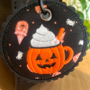 May include: A round, scalloped-edge fabric tag featuring a black background with a repeating pattern of orange jack-o'-lantern mugs topped with white whipped cream, small white ghosts, and orange accents, attached by a metal grommet and ring.