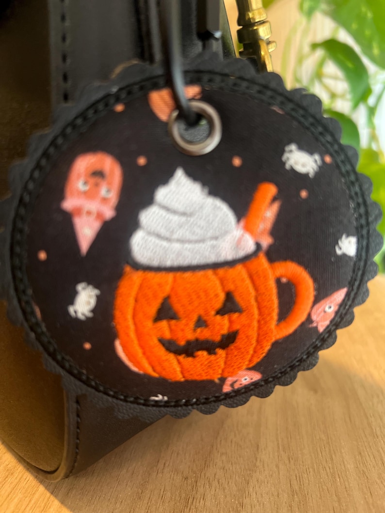May include: A round, scalloped-edge fabric tag featuring a black background with a repeating pattern of orange jack-o'-lantern mugs topped with white whipped cream, small white ghosts, and orange accents, attached by a metal grommet and ring.