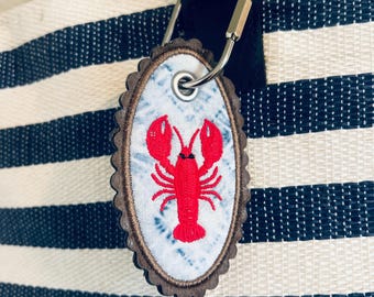 May include: An oval-shaped bag tag with a scalloped dark brown edge and a light blue tie-dye fabric center featuring a bright red embroidered lobster. The tag is attached to a black and white striped bag by a silver-toned metal carabiner clip.