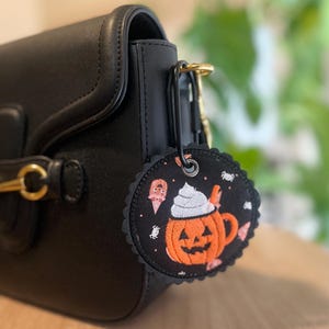 May include: A black circular bag charm with a scalloped edge, featuring an embroidered orange pumpkin mug with white whipped cream, small ghosts, and spiders. The charm is attached to a black carabiner clip hanging from a black leather handbag.