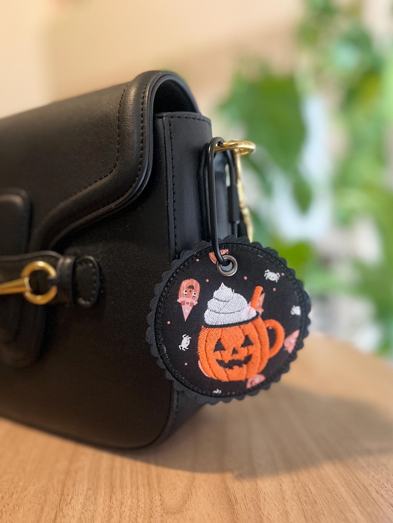 May include: A black circular bag charm with a scalloped edge, featuring an embroidered orange pumpkin mug with white whipped cream, small ghosts, and spiders. The charm is attached to a black carabiner clip hanging from a black leather handbag.