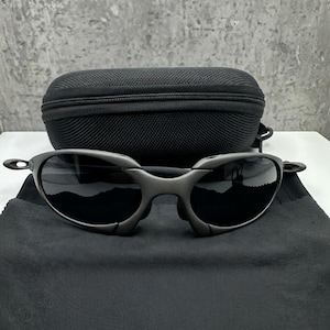 May include: Dark gray sunglasses with black lenses and a matching black case. The sunglasses are resting on a black microfiber cloth. The frame has a unique, angular design.