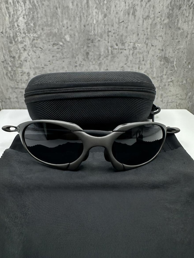 May include: Dark gray sunglasses with black lenses and a matching black case. The sunglasses are resting on a black microfiber cloth. The frame has a unique, angular design.