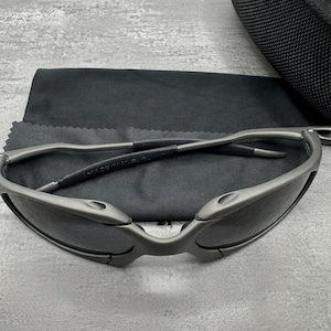 May include: Gray sunglasses with dark lenses, a sleek, modern design, and a matte finish. The sunglasses are resting on a black microfiber cloth, with a black case in the background. The frame is made of metal.