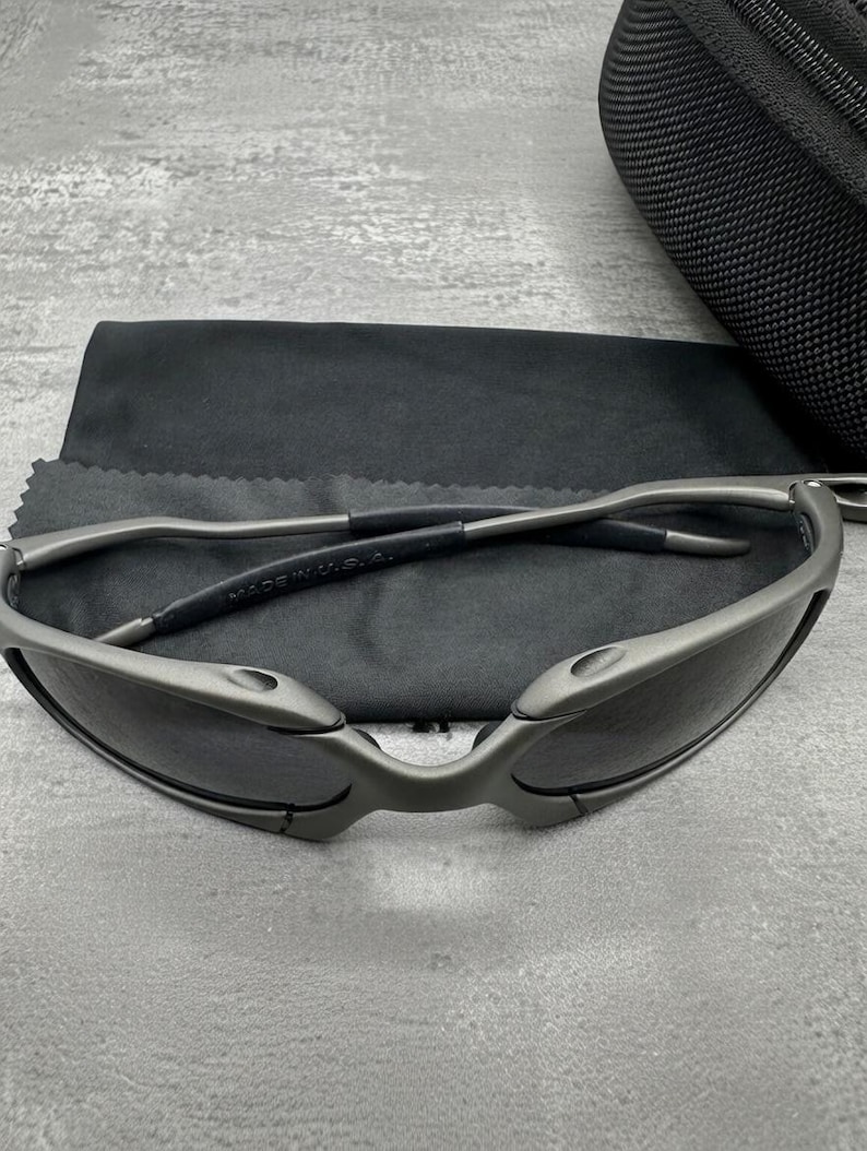 May include: Gray sunglasses with dark lenses, a sleek, modern design, and a matte finish. The sunglasses are resting on a black microfiber cloth, with a black case in the background. The frame is made of metal.