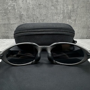 May include: Oval-shaped, dark gray sunglasses with black lenses and a matching black case. The sunglasses have a sleek, modern design, and are resting on a black surface.
