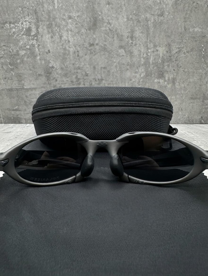 May include: Oval-shaped, dark gray sunglasses with black lenses and a matching black case. The sunglasses have a sleek, modern design, and are resting on a black surface.