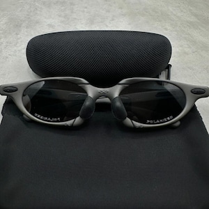 May include: Gray sunglasses with dark lenses and a matching case and pouch. The sunglasses have a curved frame and the word "POLARIZED" is visible on the lens. The case is oval-shaped and made of a textured material.