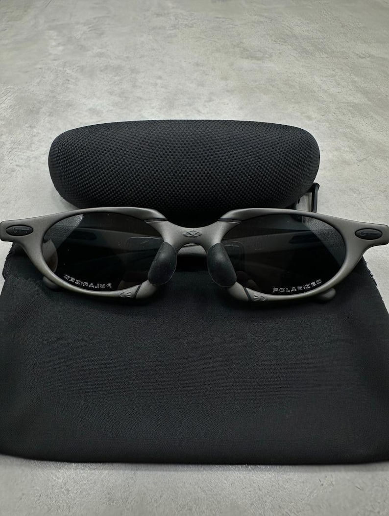 May include: Gray sunglasses with dark lenses and a matching case and pouch. The sunglasses have a curved frame and the word "POLARIZED" is visible on the lens. The case is oval-shaped and made of a textured material.