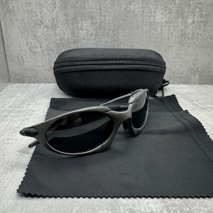 May include: Gray sunglasses with black lenses and a black carrying case. The sunglasses are on a black microfiber cloth. The case is oval-shaped and has a zipper closure. The background is a gray, textured surface.