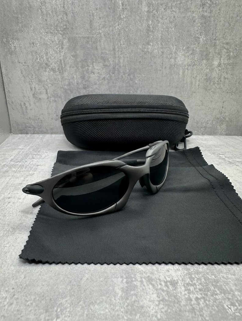 May include: Gray sunglasses with black lenses and a black carrying case. The sunglasses are on a black microfiber cloth. The case is oval-shaped and has a zipper closure. The background is a gray, textured surface.
