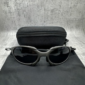 May include: Gunmetal-colored sunglasses with dark lenses rest on a black microfiber cloth. A black, zippered sunglasses case sits behind the glasses. The sunglasses have a sleek, modern design.