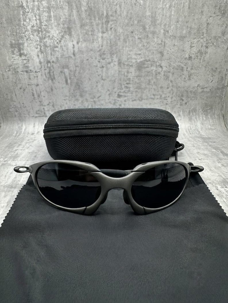 May include: Gunmetal-colored sunglasses with dark lenses rest on a black microfiber cloth. A black, zippered sunglasses case sits behind the glasses. The sunglasses have a sleek, modern design.